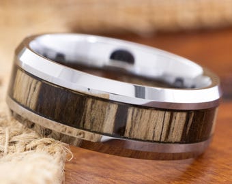 May include: A silver-toned ring with a wood-grain inlay. The ring features a polished silver band and a dark brown and tan wood-grain pattern around the center. The ring is set against a wooden surface.