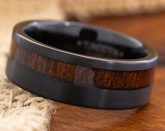 May include: Black tungsten ring with a dark wood inlay. The ring features a polished black band with a contrasting band of rich, dark brown wood. The wood grain is visible, adding texture to the ring's design. A modern and stylish piece of jewelry.