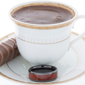 Op de afbeelding: Een wit koffiekopje met een gouden rand en een schoteltje, gevuld met donkere koffie. Een chocolade reep en een ring van hout en zwart liggen op het schoteltje.