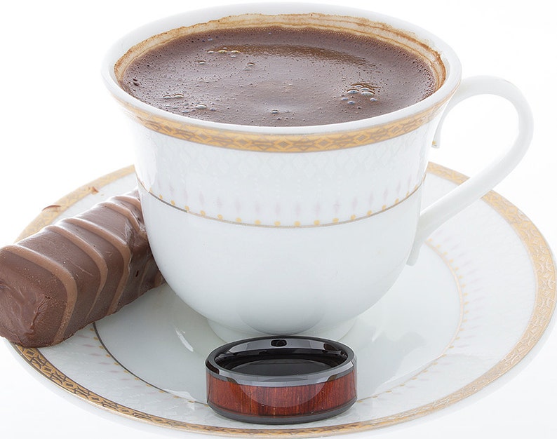 Op de afbeelding: Een wit koffiekopje met een gouden rand en een schoteltje, gevuld met donkere koffie. Een chocolade reep en een ring van hout en zwart liggen op het schoteltje.
