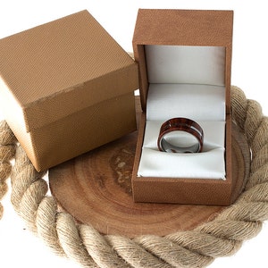 May include: A brown wooden ring box with a white satin interior. A dark brown and silver ring with a wood inlay is displayed inside the box.