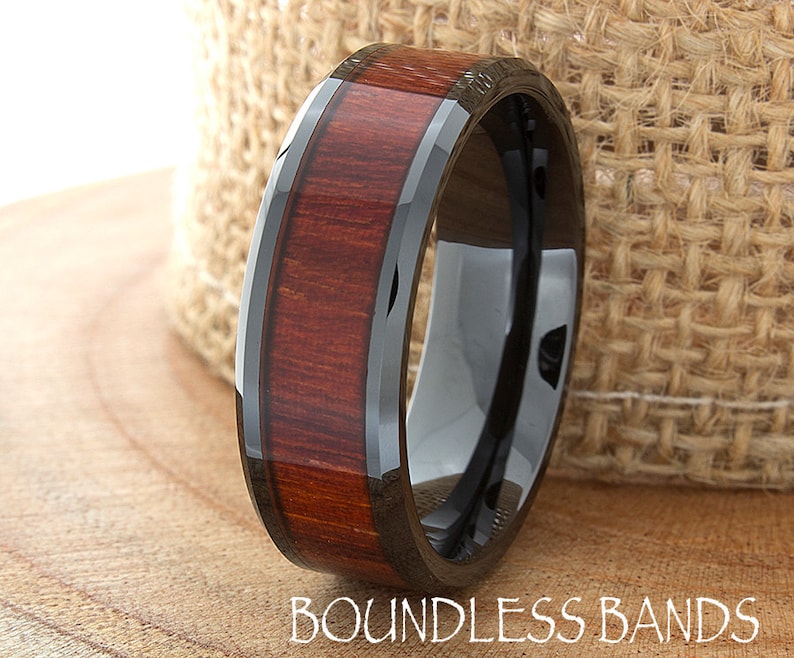 Op de afbeelding: Een zwarte keramische ring met een band van roodbruin houtinleg. De ring heeft een afgeschuinde rand en is gepolijst tot een hoogglans. "Boundless Bands" is in wit gedrukt op de onderkant van de afbeelding.