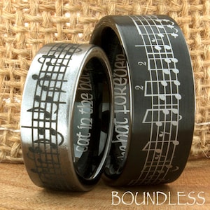 May include: Two black tungsten carbide wedding bands with a musical note design. The band on the left has the inscription "Got in to the beat". The band on the right has the inscription "In that for good".