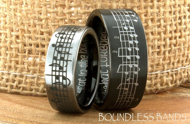 May include: Two black tungsten carbide wedding bands with a musical note design. The band on the left has the inscription "Got in to the beat". The band on the right has the inscription "In that for good".