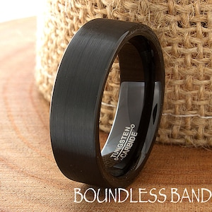 Op de afbeelding: Een zwarte ring met een geborstelde afwerking en een plat profiel. De binnenkant van de band is voorzien van de gegraveerde tekst TUNGSTEN CARBIDE. De ring staat rechtop en toont het donkere metalen oppervlak en de duidelijke inscriptie.