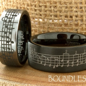 May include: Two black tungsten carbide rings with a musical note design. The ring on the left has the words "Got it in the bag" engraved on the side. The ring on the right has a musical staff with notes and numbers engraved on the side.