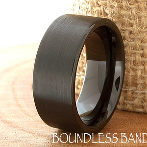Puede incluir: Un anillo de carburo de tungsteno negro cepillado con un borde biselado. El anillo se muestra sobre una superficie de madera.