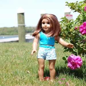 May include: A doll wearing a blue and white patterned outfit stands in a grassy area with pink flowers in the background.