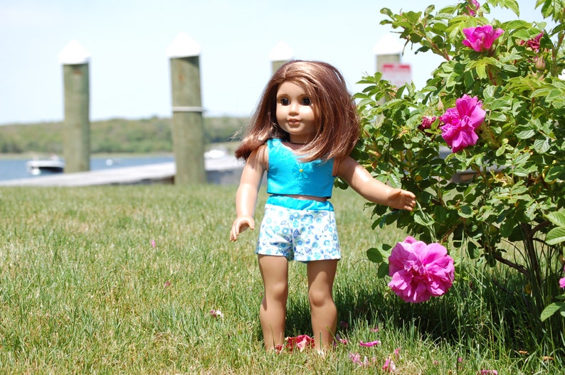 May include: A doll wearing a blue and white patterned outfit stands in a grassy area with pink flowers in the background.