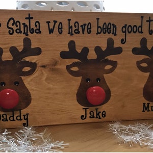 May include: Wooden sign with three reindeer silhouettes, each with a red nose. The sign reads "Dear Santa we have been good this year" and has the names "Daddy", "Jake", and "Mummy" written below the reindeer.