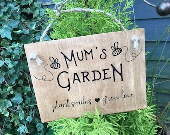 Könnte beinhalten: Rechteckiges Holzschild mit dem schwarzen Schriftzug "Mum's Garden", verziert mit Bienenmotiven. Das Schild enthält auch die Sätze "plant smiles" und "grow love". Es wird an einem Seil aufgehängt.