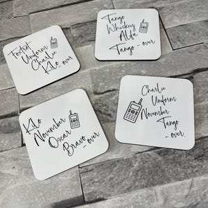 Peut inclure: Quatre sous-verres carrés blancs avec du texte noir et une petite illustration noire d'une talkie-walkie sur chacun. Le texte sur les sous-verres est : "Foxtrot Uniform Charlie Kilo - over", "Kilo November Oscar Bravo - over", "Tango Whiskey Alfa Tango - over", et "Charlie Uniform November Tango - over".