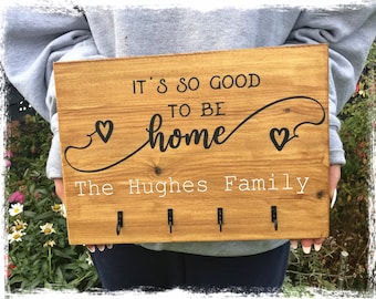 Puede incluir: Porta llaves de madera con el texto "It's so good to be home" y "The Hughes Family" con dos símbolos de corazón y cuatro ganchos.