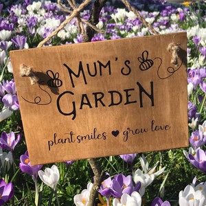 Może przedstawiać: Drewniany znak z napisem "Mum's Garden" i frazą "plant smiles grow love". Znak wisi w ogrodzie z fioletowymi i białymi krokusami.