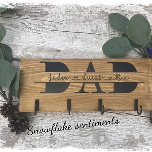 May include: Wooden key holder with the word "DAD" in large black letters and the names "Jaden", "Lucas", and "Ben" written below in a smaller font. The key holder has four hooks and the words "Snowflake Sentiments" are written below the hooks.