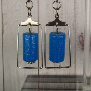 May include: A pair of silver earrings with a blue, textured, cylindrical bead hanging from a rectangular frame.