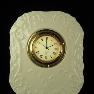 May include: A white porcelain clock with a gold rim and Roman numeral face. The clock is decorated with a floral design and reads "LENOX QUARTZ".