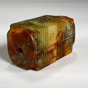 Puede incluir: Un colgante de jade tallado con forma rectangular y un agujero en el centro. El jade es una mezcla de verde, marrón y blanco.