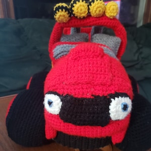 May include: A handmade crochet toy car in red, black, yellow, white, and gray. The car features large black tires, a red body, and a black front with white and blue eyes. The top has yellow and black pom-poms.