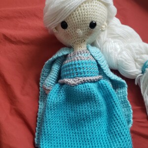 May include: A handmade crochet doll with white yarn hair styled in a braid and a blue dress. The doll has black button eyes and a light blue cape. The dress has a gray and silver trim around the waist.