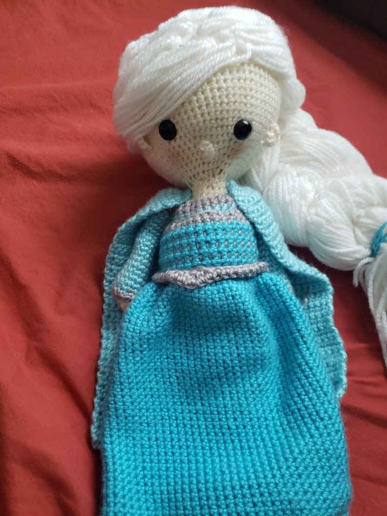 May include: A handmade crochet doll with white yarn hair styled in a braid and a blue dress. The doll has black button eyes and a light blue cape. The dress has a gray and silver trim around the waist.