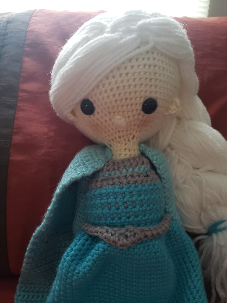 May include: A handmade crochet doll with white hair styled in a braid and long flowing locks. The doll wears a blue dress with a gray bodice and a matching cape. Black button eyes and rosy cheeks complete the design.