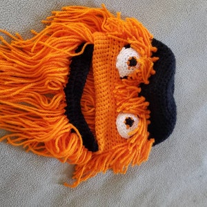 May include: A handmade, orange and black crochet hat shaped like a cartoon dog. The hat features large white eyes with black pupils and long, orange yarn strands for fur.