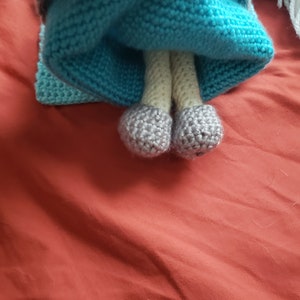May include: A handmade doll with a blue knitted dress and gray shoes. The doll's legs are beige, and the dress has a textured appearance. The doll is on a red surface.