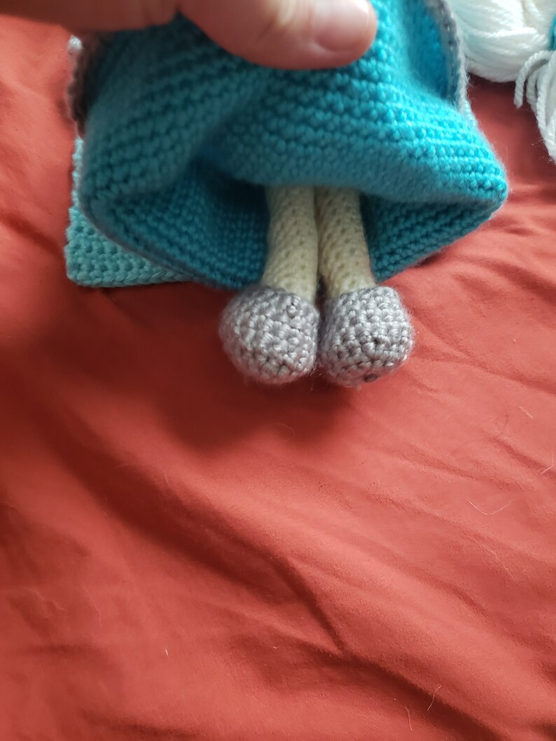 May include: A handmade doll with a blue knitted dress and gray shoes. The doll's legs are beige, and the dress has a textured appearance. The doll is on a red surface.