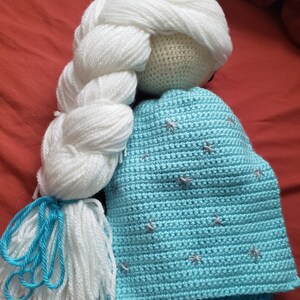 May include: A handmade crochet doll with a white yarn braid and a light blue dress. The doll's dress is adorned with embroidered star details. The doll's head is a light beige color, and the braid is tied with blue yarn.