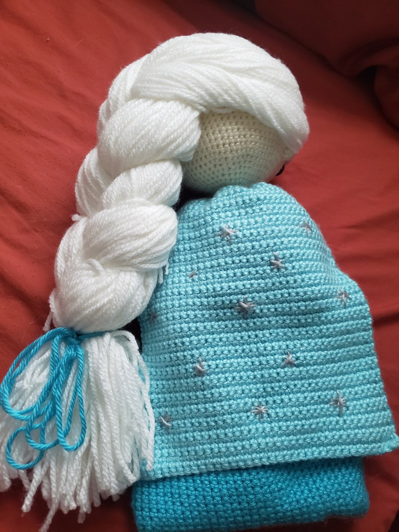 May include: A handmade crochet doll with a white yarn braid and a light blue dress. The doll's dress is adorned with embroidered star details. The doll's head is a light beige color, and the braid is tied with blue yarn.