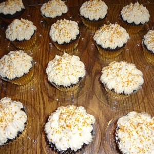 May include: A close-up of a tray of chocolate cupcakes with white icing and gold sprinkles. The cupcakes are arranged in rows and are in clear plastic containers.