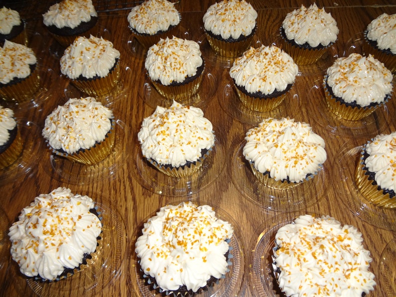 May include: A close-up of a tray of chocolate cupcakes with white icing and gold sprinkles. The cupcakes are arranged in rows and are in clear plastic containers.