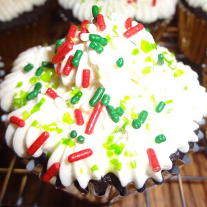 May include: A close-up of a chocolate cupcake with white icing and green and red sprinkles. The cupcake is on a wire rack.