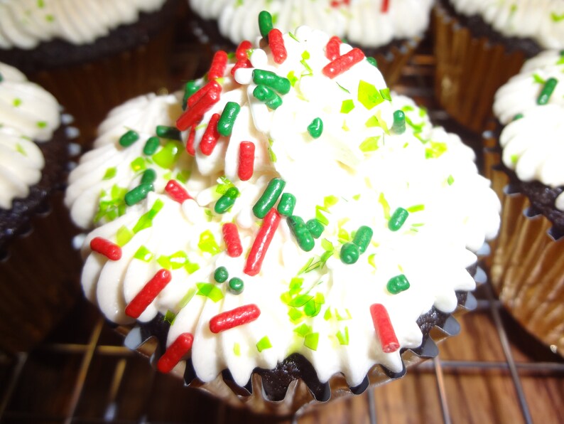 May include: A close-up of a chocolate cupcake with white icing and green and red sprinkles. The cupcake is on a wire rack.