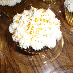 May include: A close-up of a chocolate cupcake topped with white icing and gold sprinkles. The cupcake is in a gold foil liner and is sitting on a clear plastic tray.