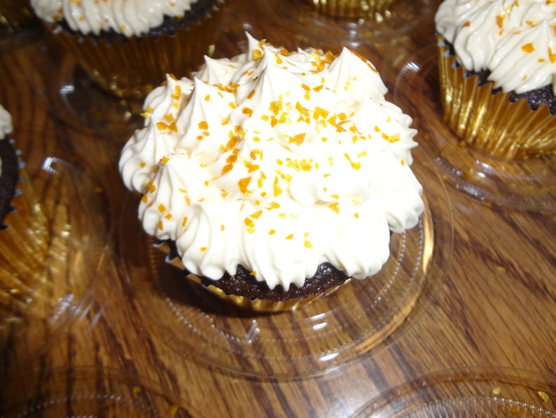 May include: A close-up of a chocolate cupcake topped with white icing and gold sprinkles. The cupcake is in a gold foil liner and is sitting on a clear plastic tray.