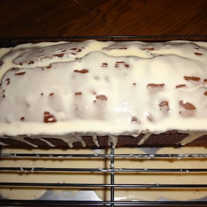 May include: A loaf of chocolate bread topped with a white glaze. The glaze is dripping down the sides of the bread onto a wire rack.