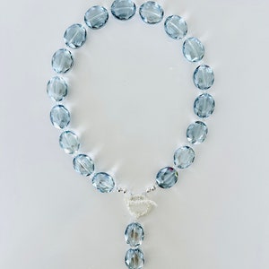 Könnte beinhalten: Eine hellblaue Kristallkette mit einem silbernen Anhänger. Die Kette besteht aus ovalen Perlen und der Anhänger ist ein gedrehtes Silberdesign.