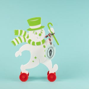 May include: A whimsical snowman figure with a green top hat, scarf, and cane. The snowman is white with green accents and red wheels. It holds a tag with the text "Witch House Design". The background is a light blue.