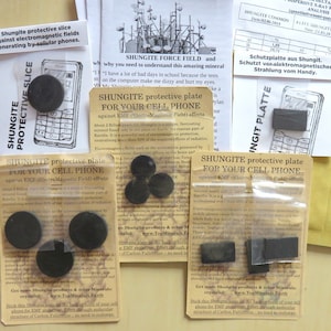以下が含まれることがあります： 透明なプラスチック袋に詰められた、小さな黒いシュンガイト保護プレート3枚のクローズアップ。プレートには、「携帯電話用シュンガイト保護プレート EMF（電磁場）の影響から保護」とラベルが貼られています。