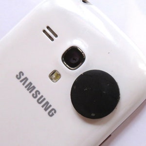 May include: White Samsung smartphone with a black round sticker on the back.