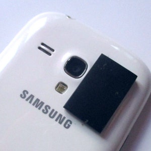May include: A white Samsung smartphone with a black square object covering the camera lens. The Samsung logo is visible on the back of the phone.