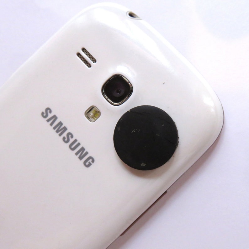 May include: White Samsung smartphone with a black round sticker on the back.