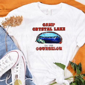 May include: White t-shirt with a graphic that reads "Camp Crystal Lake Counselor" in red text. The graphic includes an image of a lake scene. The shirt is styled with blue jeans, white sneakers, and an orange sweater.