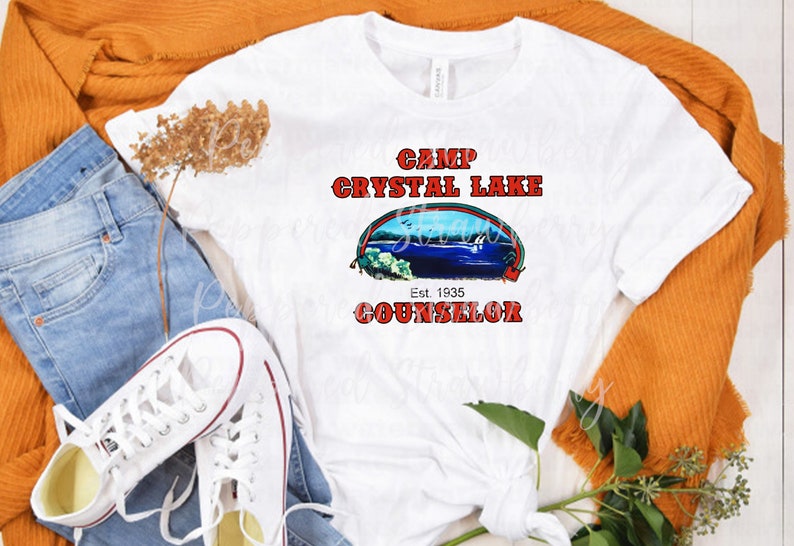 May include: White t-shirt with a graphic that reads "Camp Crystal Lake Counselor" in red text. The graphic includes an image of a lake scene. The shirt is styled with blue jeans, white sneakers, and an orange sweater.