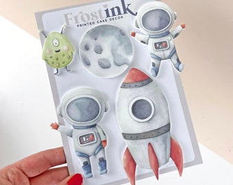 May include: A set of five printable cake decorations featuring a rocket, two astronauts, a moon, and a green monster. The decorations are printed on white cardstock and are perfect for adding a space theme to your next cake.