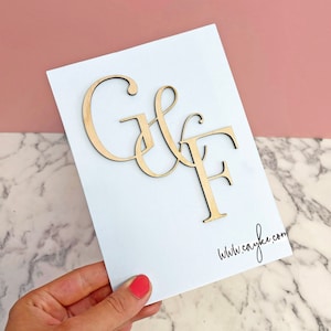 May include: A white card with the letters "G & F" in cursive, cut out of wood and attached to the card. The letters are in a light brown colour. The text "www.cayflee.com" is printed in black at the bottom right corner of the card.