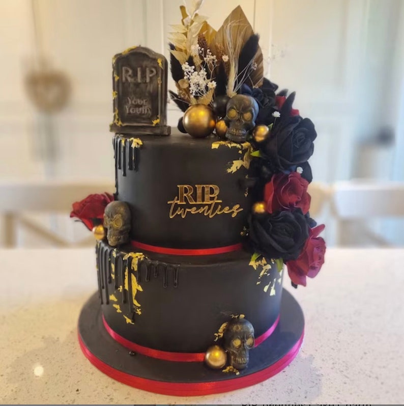 May include: A two-tiered black cake decorated with gold leaf, red and black roses, and skulls. The cake is topped with a tombstone that reads "RIP Your Youth". The bottom tier of the cake has the words "RIP Twenties" written in gold.