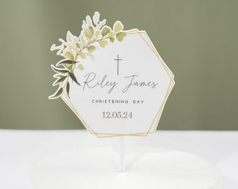 May include: White cake topper with a gold hexagonal frame and green floral design. The text reads "Riley James" and "Christening Day" with the date "12.05.24".